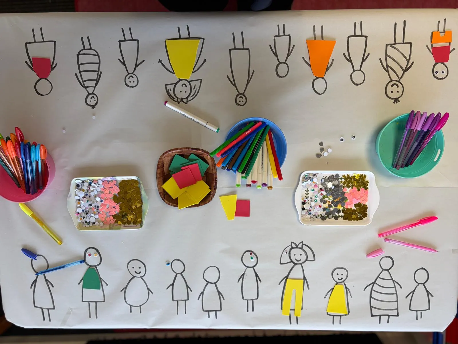 A craft table set up with pens, sequins and paper people drawn by tamariki
